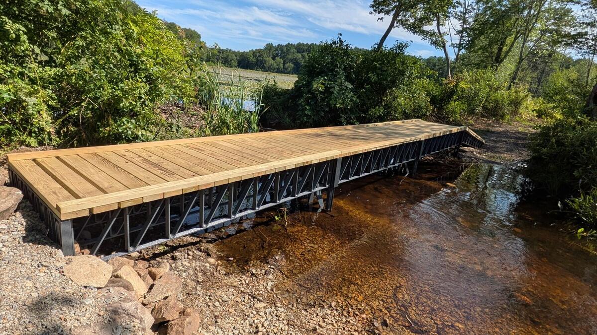 A finished footbridge with a wooden deck on black composite trusses, crossing a shallow stream