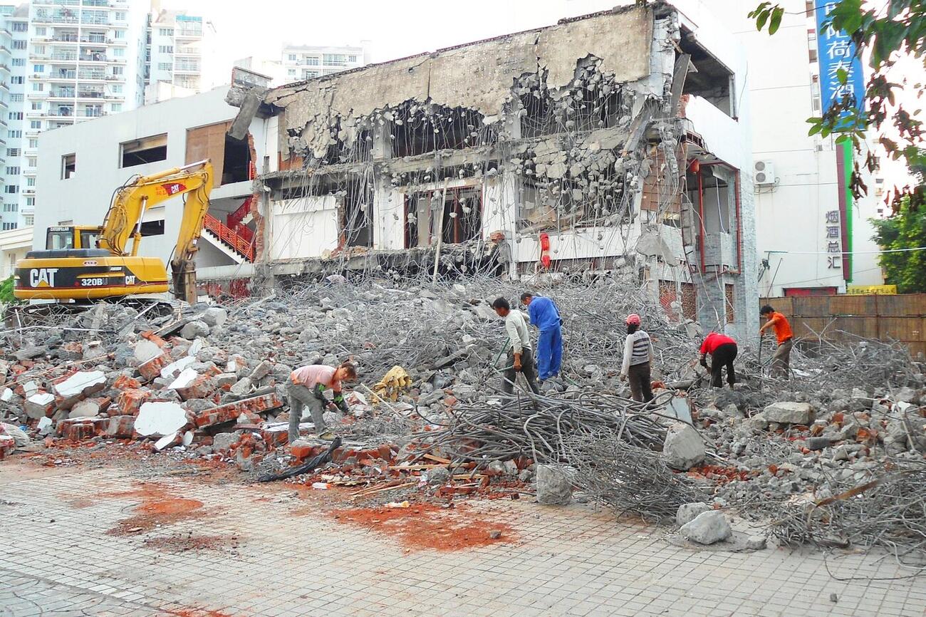 Workers pick through concrete rubble and rebar beside a partly demolished building, with an excavator standing by